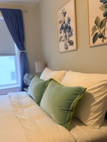A bedroom with white bedding, green pillows and botanical prints
