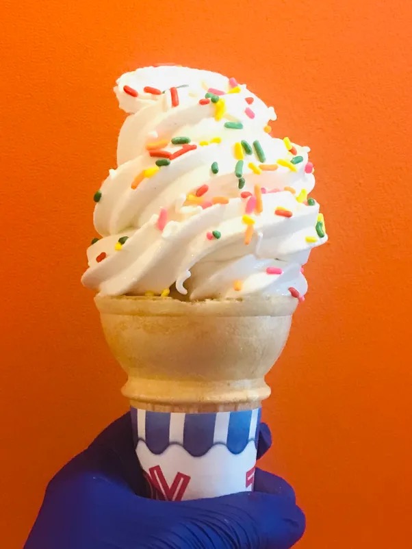 A soft serve cone with rainbow sprinkles held up against an orange wall